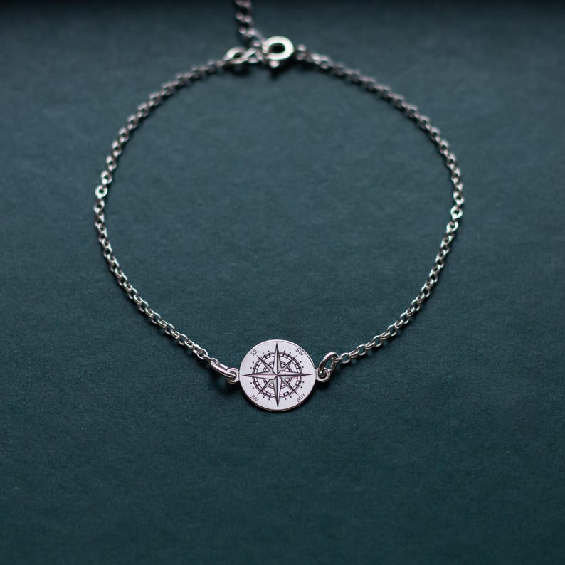 Puede incluir: Una pulsera de cadena plateada con un dije circular en el centro. El dije est&aacute; grabado con un dise&ntilde;o detallado de rosa de los vientos, incluyendo los puntos cardinales N, S, E y O. La pulsera incluye un peque&ntilde;o cierre de mosquet&oacute;n.