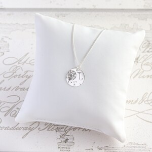May include: A silver necklace with a round pendant featuring a dandelion design. The pendant is suspended from a delicate chain and rests on a white pillow. The background is a light-colored surface with script.