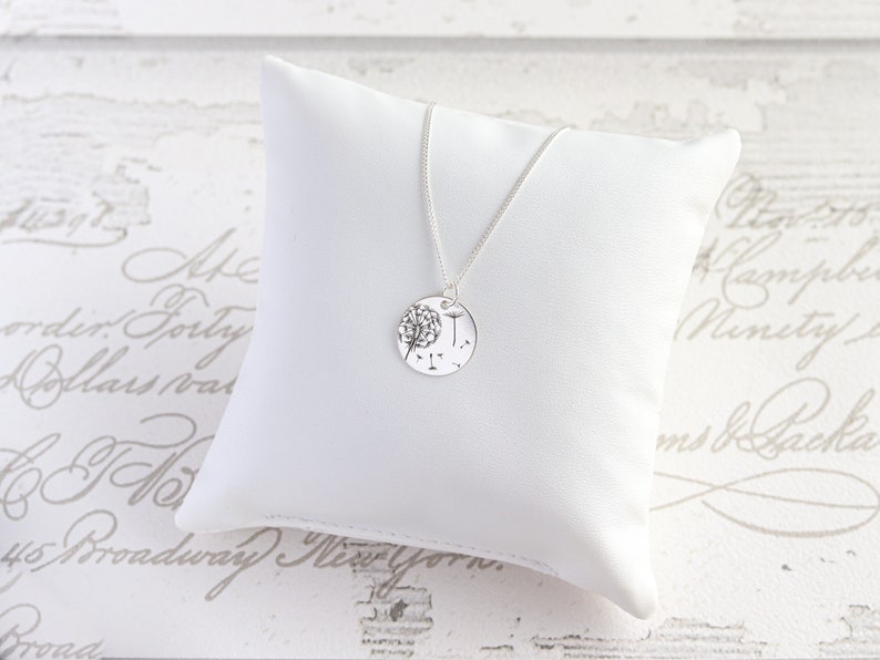 May include: A silver necklace with a round pendant featuring a dandelion design. The pendant is suspended from a delicate chain and rests on a white pillow. The background is a light-colored surface with script.