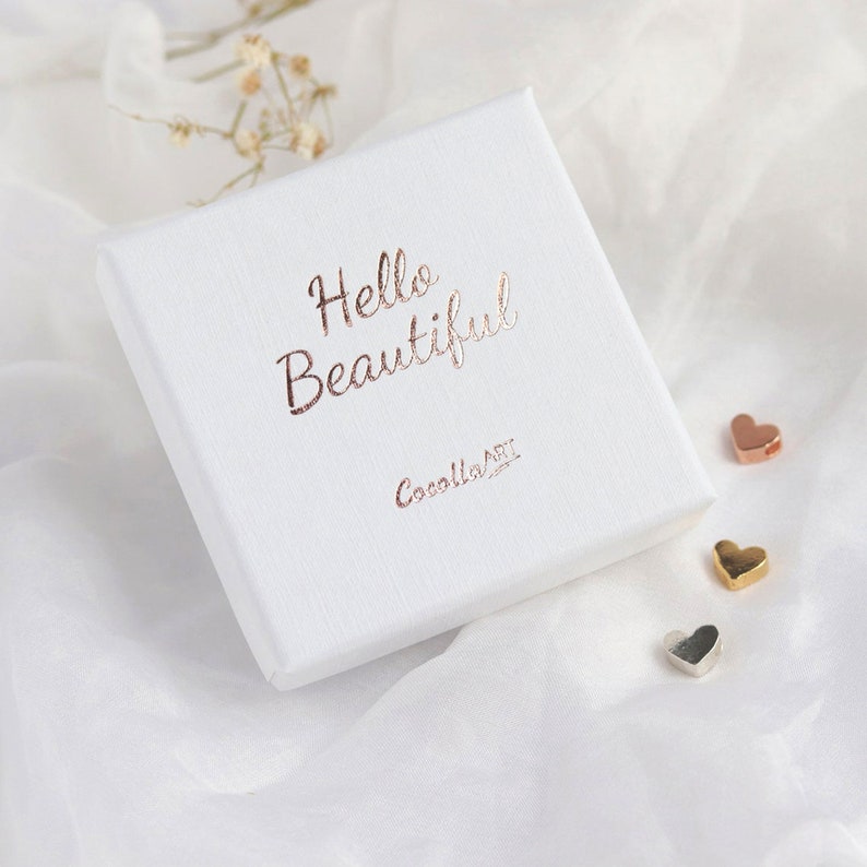 Puede incluir: Una caja de regalo cuadrada blanca con superficie texturizada que presenta el texto Hello Beautiful en l&aacute;mina de oro rosa y el nombre de la marca CocollaART. Tres peque&ntilde;os dijes en forma de coraz&oacute;n en tonos oro rosa, oro y plata junto a la caja.