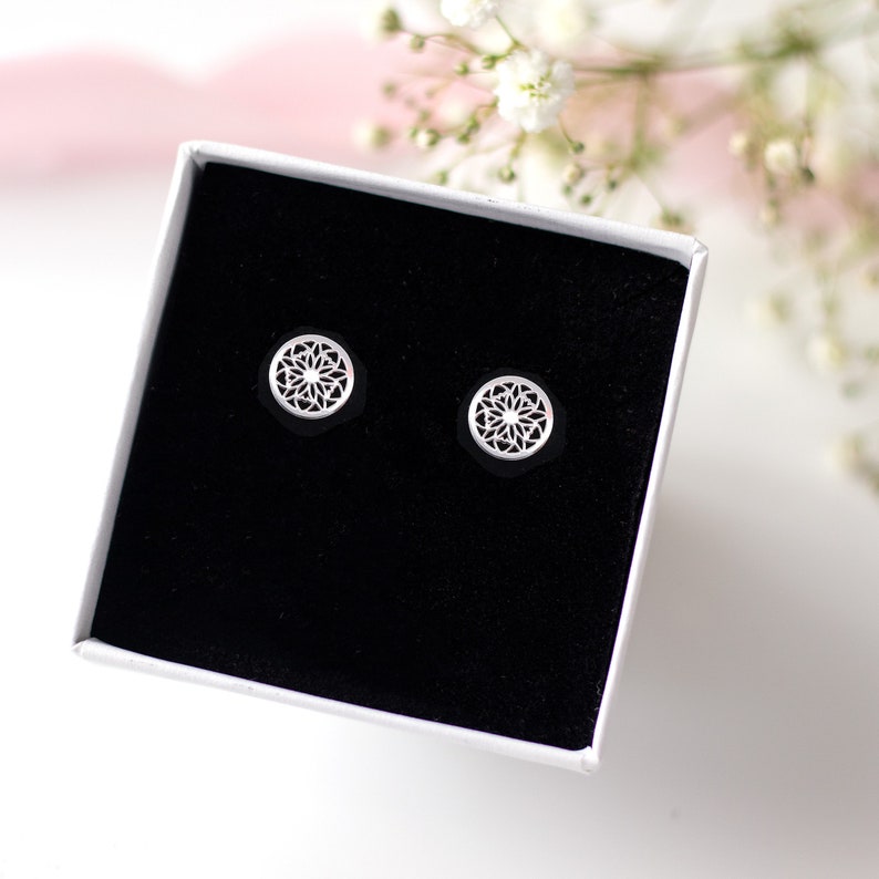 A pair of sterling silver stud earrings with intricate geometric patterns displayed in a black velvet box with a soft pink and white floral background.