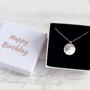 May include: A silver necklace with a round pendant featuring a dandelion design, presented in a white gift box. The box is open, revealing the necklace against a black interior. The box lid has "Happy Birthday" in a copper color.
