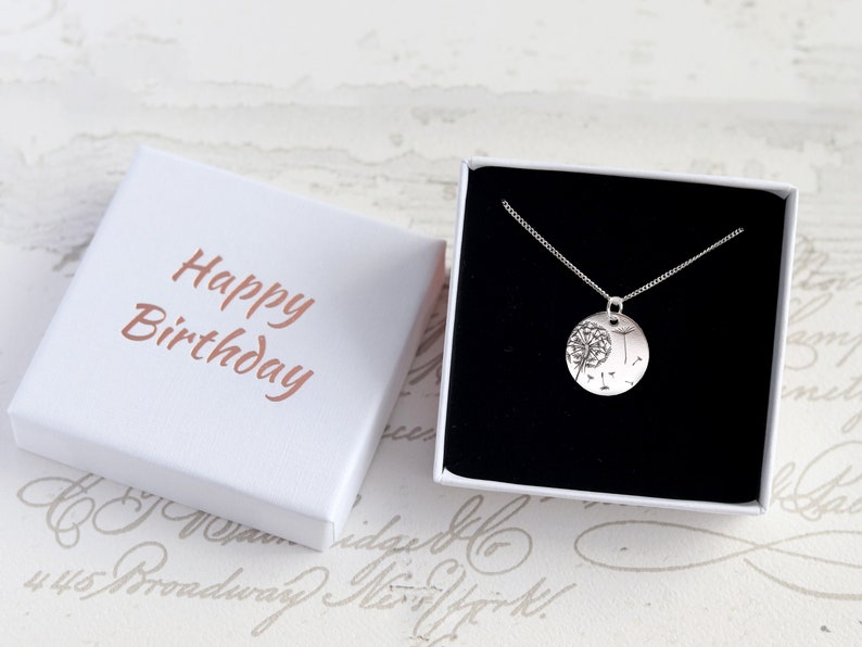 May include: A silver necklace with a round pendant featuring a dandelion design, presented in a white gift box. The box is open, revealing the necklace against a black interior. The box lid has "Happy Birthday" in a copper color.