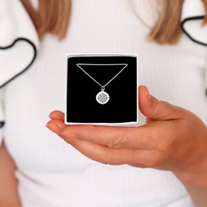 Op de afbeelding: Een zilverkleurige kompas-hangerketting gepresenteerd in een witte geschenkdoos met een zwart interieur. De kompas-hanger is rond met gedetailleerde markeringen. De ketting is delicaat. De doos wordt vastgehouden door een persoon die een witte top draagt.