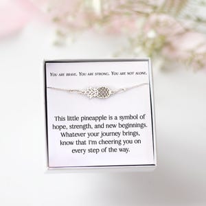 Peut inclure: Un bracelet à breloque ananas argenté présenté dans une boîte blanche. La carte à l'intérieur de la boîte indique : "YOU ARE BRAVE. YOU ARE STRONG. YOU ARE NOT ALONE." et "This little pineapple is a symbol of hope, strength, and new beginnings."