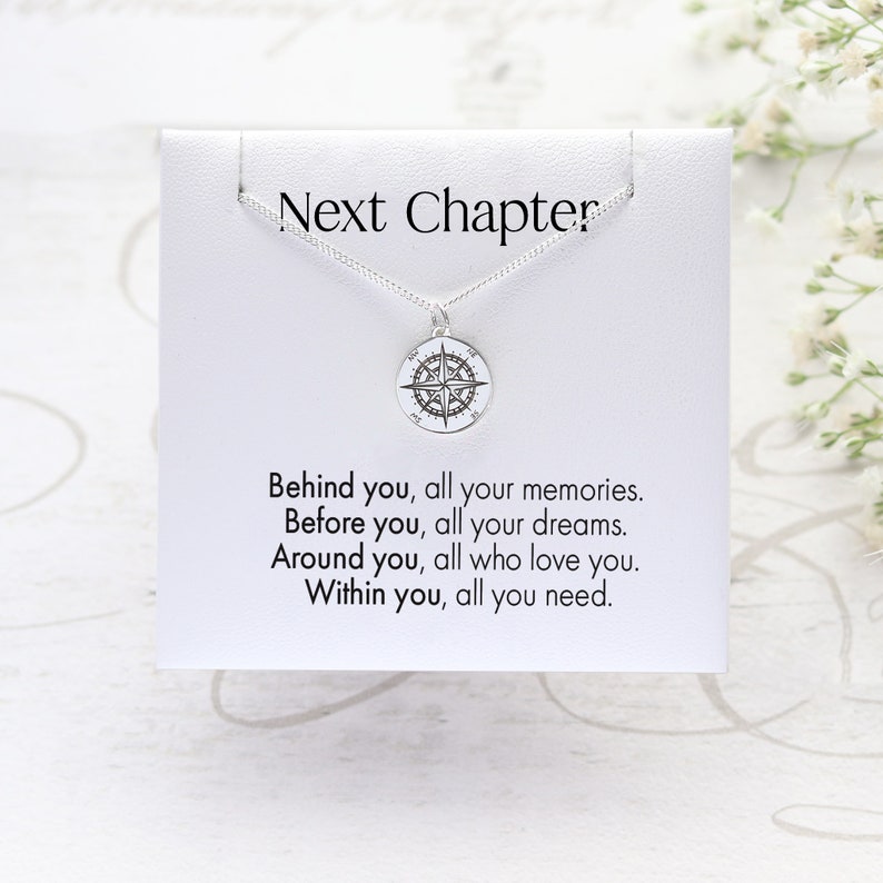 Op de afbeelding: Een zilveren kompas hanger ketting op een witte kaart. De kaart heeft de tekst "Next Chapter" boven de ketting en een motiverende quote eronder. De ketting is een ronde hanger met een kompas ontwerp.