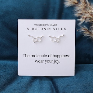 May include: A pair of sterling silver Serotonin Studs earrings displayed on a white card. The card reads "925 Sterling Silver Serotonin Studs" and "The molecule of happiness. Wear your joy."