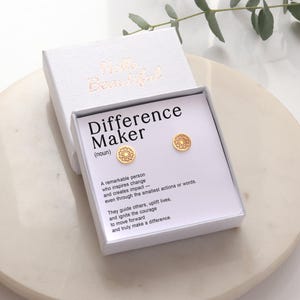 May include: A white gift box with gold lettering reading "Hello Beautiful". Inside, a pair of gold stud earrings with an openwork circular design is presented on a card that reads "Difference Maker". The earrings are on a white background.