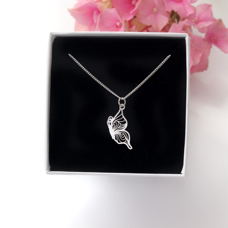 Pode incluir: Um colar com pingente de borboleta prateada exibido em uma caixa de presente branca com interior preto. O pingente de borboleta apresenta um design intrincado e delicado com padr&otilde;es vazados. Flores cor-de-rosa vis&iacute;veis no fundo.