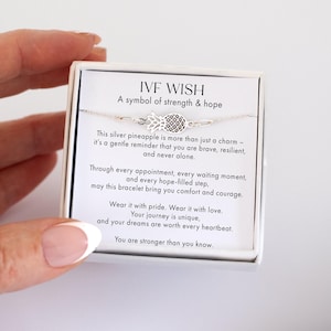 May include: A silver pineapple charm bracelet presented in a white box. The box has text that reads "IVF WISH, A symbol of strength & hope." The bracelet is delicate and the pineapple charm is detailed.