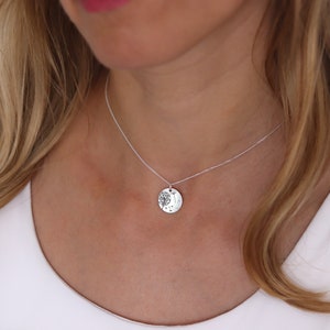 May include: A silver necklace with a round pendant featuring a tree design. The pendant hangs from a delicate chain. The necklace is worn against a white top.