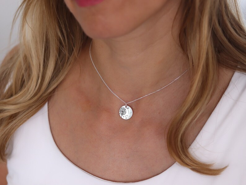 May include: A silver necklace with a round pendant featuring a tree design. The pendant hangs from a delicate chain. The necklace is worn against a white top.