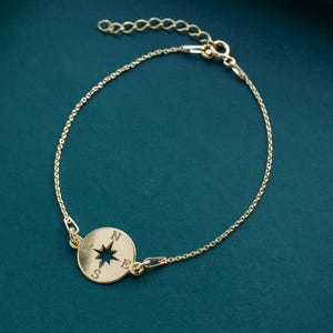 May include: A gold bracelet with a compass pendant. The bracelet has a fine chain and a circular charm with the cardinal points (N, S, E, W) and a star. The bracelet is shown against a teal background.