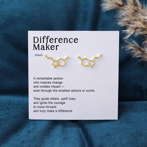 May include: A pair of gold-colored stud earrings in the shape of a serotonin molecule, displayed on a white card with the text "Difference Maker". The card includes a definition and inspirational message.