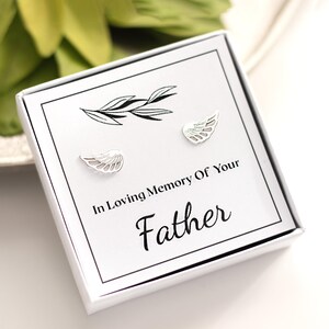 Puede incluir: Una caja de regalo blanca con pendientes plateados en forma de alas. La caja tiene el texto "In Loving Memory Of Your Father" en escritura negra. Una ramita decorativa está impresa encima de los pendientes.