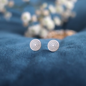 Könnte beinhalten: Ein Paar silberfarbene Ohrstecker. Jeder Ohrstecker hat eine runde Form mit einem detaillierten, durchbrochenen floralen Design. Die Ohrringe werden auf einer teal-farbenen Samtoberfläche mit weichen, verschwommenen floralen Akzenten im Hintergrund präsentiert.