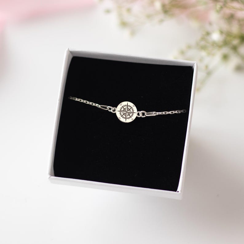 Puede incluir: Una pulsera de cadena en tono plateado con un dije circular grabado con un dise&ntilde;o de rosa de los vientos en negro. La pulsera se presenta dentro de una caja de regalo cuadrada blanca con un inserto de terciopelo negro, mostrando los eslabones.
