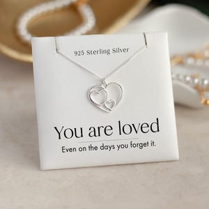 Puede incluir: Un collar de plata de ley 925 con un colgante de tres corazones entrelazados. La joya se presenta sobre una tarjeta blanca con el texto impreso "925 Sterling Silver", "You are loved" y "Even on the days you forget it."