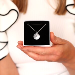 May include: A silver necklace with a round pendant featuring a tree design, presented in a white box with a black interior. The necklace is on a delicate chain. The box is held in a person's hands.