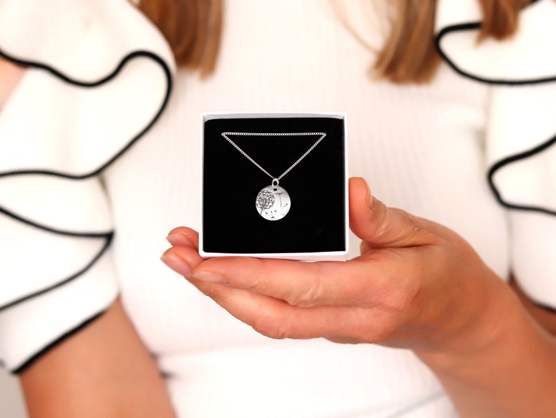 May include: A silver necklace with a round pendant featuring a tree design, presented in a white box with a black interior. The necklace is on a delicate chain. The box is held in a person's hands.