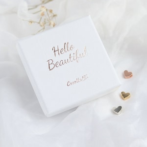 Pode incluir: Uma caixa quadrada branca com as palavras "Hello Beautiful" em escrita em ouro rosa. A caixa tamb&eacute;m tem o texto "Cocalla ART" em ouro rosa. Tr&ecirc;s pequenos pingentes em forma de cora&ccedil;&atilde;o em ouro rosa, ouro e prata est&atilde;o ao lado da caixa.
