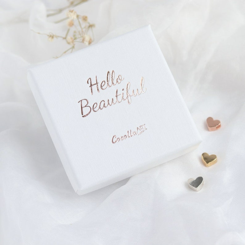 Pode incluir: Uma caixa quadrada branca com as palavras "Hello Beautiful" em escrita em ouro rosa. A caixa tamb&eacute;m tem o texto "Cocalla ART" em ouro rosa. Tr&ecirc;s pequenos pingentes em forma de cora&ccedil;&atilde;o em ouro rosa, ouro e prata est&atilde;o ao lado da caixa.