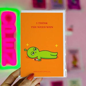 Könnte beinhalten: Orangefarbene Grußkarte mit dem Text "I THINK THE WEED WON" und einem Cartoon-Alien, das eine Zigarette raucht. Im Hintergrund ist ein grünes Neon-"OPEN"-Schild zu sehen.