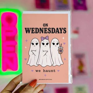 Peut inclure: Carte de vœux avec un fond pêche représentant trois fantômes de dessins animés avec le texte "ON WEDNESDAYS we haunt". Un fantôme tient une tasse de café. La carte est dans une pochette en plastique, avec une enseigne "OPEN" rose néon en arrière-plan.
