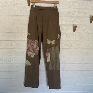 Puede incluir: Pantalón marrón con un diseño de patchwork con motivos florales y mariposas. El pantalón tiene una pierna recta y un ajuste holgado.