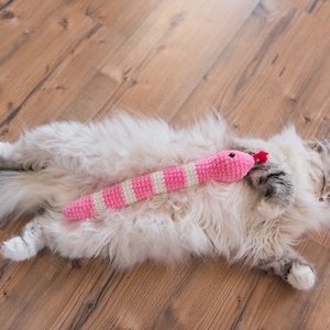 May include: A fluffy white cat with a long tail is lying on a wooden floor. The cat is holding a pink and white crocheted snake toy in its paws.