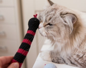 May include: A fluffy white cat with long fur is sniffing a red and black crocheted toy snake.