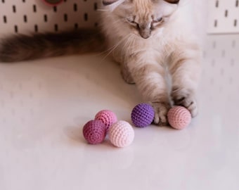 May include: A white cat playing with five crocheted balls in shades of pink and purple. The balls are on a white surface.