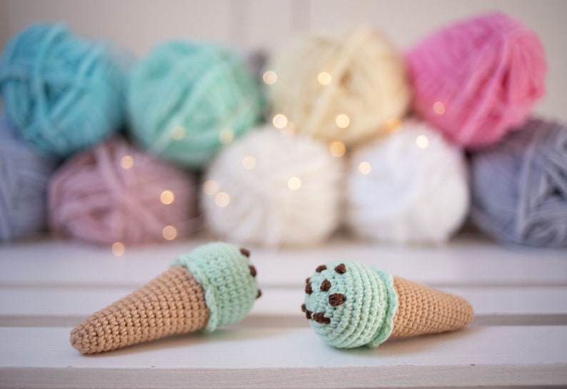 Puede incluir: Dos conos de helado de ganchillo, uno con helado de menta verde y el otro con helado de chocolate con chispas. Los conos est&aacute;n sobre una superficie blanca.