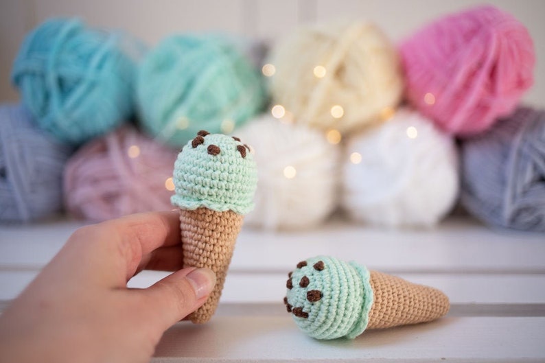 Puede incluir: Dos conos de helado de crochet con helado verde menta y virutas de chocolate. Los conos est&aacute;n sobre una superficie blanca.