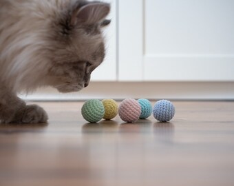 May include: A fluffy gray cat looks at a row of five crocheted balls in pastel colors. The balls are light green, yellow, pink, blue, and light blue.