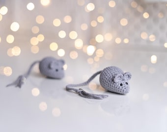 May include: Two gray crocheted mice toys with long tails. The mice are on a white surface with a blurred background of white lights.