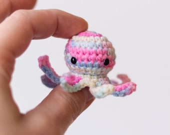 May include: A small crocheted octopus with pink, blue, and white stripes. The octopus has black eyes and is held in a hand.