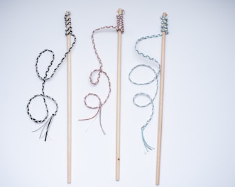 Può includere: Tre giocattoli per gatti in legno con corde di diversi colori. Le corde sono legate alla parte superiore dei bastoncini di legno e sono annodate e attorcigliate. Le corde sono nere e bianche, marroni e bianche e blu e bianche.