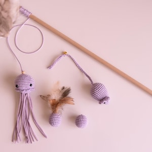 Può includere: Un set di giocattoli per gatti in crochet viola, tra cui una medusa, un topo e due palline, attaccati a una bacchetta di legno.