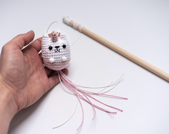 May include: A white crocheted cat toy with pink accents and a pink unicorn horn. The toy has a long tail with white and pink strings attached to a wooden stick.
