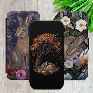 May include: Three rectangular phone cases featuring rabbit illustrations. The left case shows a rabbit among purple flowers. The centre case depicts a sleeping rabbit under an autumn tree with orange leaves. The right case shows a rabbit resting in white flowers.