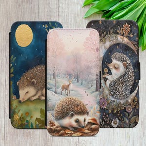 May include: Three phone cases with hedgehog designs. One shows a sleeping hedgehog under a golden moon against a dark blue night sky. Another depicts a hedgehog in a snowy forest with a deer. The third features a hedgehog on a crescent moon.