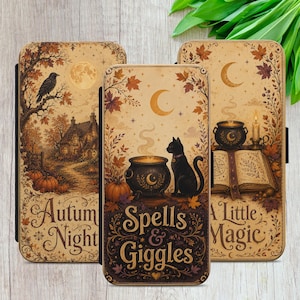 May include: Three rectangular phone cases featuring autumn-themed illustrations. The left case shows a house, a raven, and the text Autumn Night. The centre case depicts a black cat, a cauldron, and the text Spells & Giggles. The right case displays a book, a candle, and the text A Little Magic.