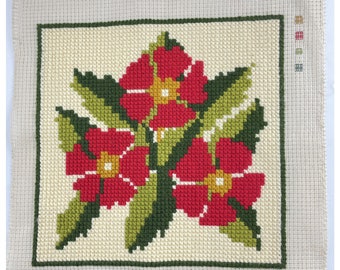 May include: Cross-stitch floral design featuring three red flowers with yellow centers and green leaves, set against a cream-colored background. The design is framed by a green border and is on a beige canvas.