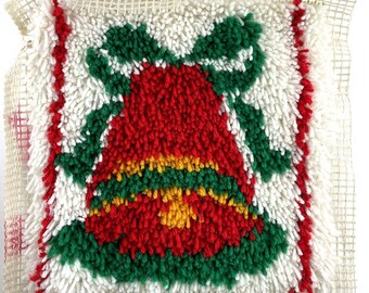 May include: A square, handmade Christmas pillow with a red bell design, green bow, and yellow accents. The bell is surrounded by green leaves and red trim. The background is white with a mesh backing.