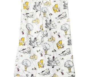 May include: White fabric baby bib with a repeating pattern of Winnie the Pooh characters, including Pooh Bear, Eeyore, Piglet, and Tigger. The bib is hanging from a wooden clothespin and twine. The bib has tie closures at the top.