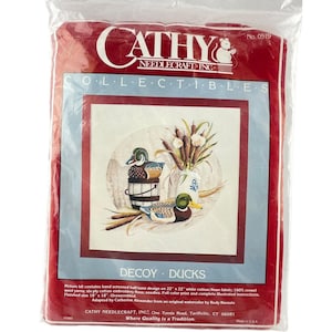 Puede incluir: Un paquete de plástico rojo que contiene un kit de punto de cruz Cathy Needlecraft Inc. Decoy Ducks. El kit presenta un diseño de dos patos, juncos y un jarrón. El paquete incluye el texto "Collectibles" y "Decoy Ducks".