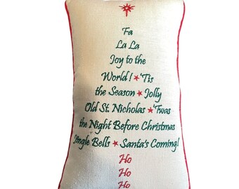 May include: Rectangular Christmas pillow with a cream-colored background and red trim. The pillow features embroidered text in green and red, including phrases like "Fa La La" and "Jingle Bells." A star design is at the top.
