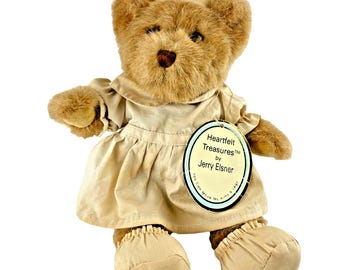 May include: A brown plush teddy bear dressed as a nurse, wearing a beige uniform with a matching cap featuring a red cross. An oval tag attached to the dress reads Heartfelt Treasures by Jerry Elsner and You Can Move My Arms & Legs.
