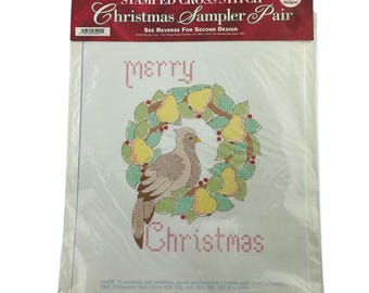 May include: A Bucilla stamped cross stitch Christmas Sampler Pair kit. The design features a dove in a wreath with pears and the words "Merry Christmas" in cross-stitch. The kit includes two designs.